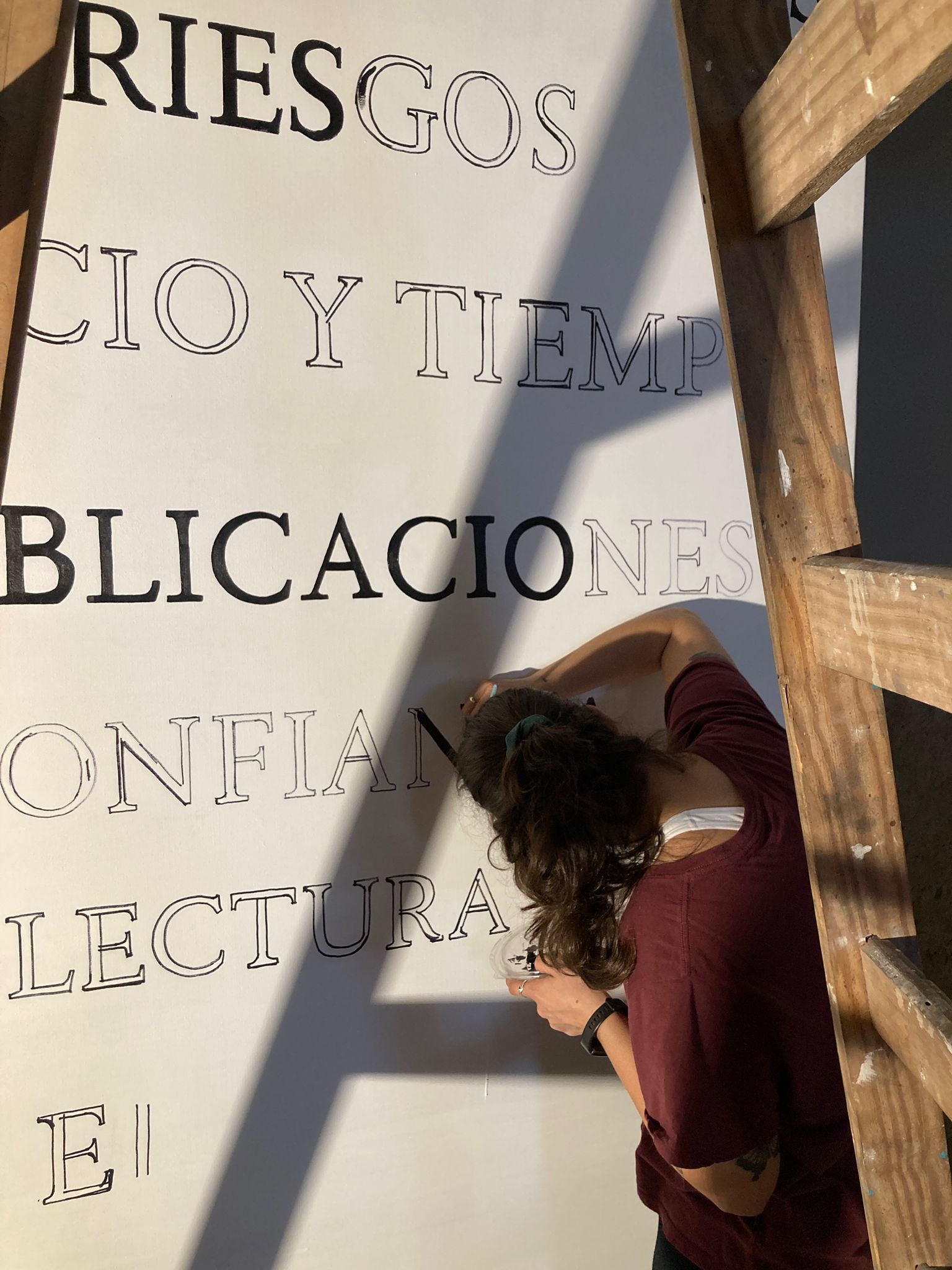 Fotografía donde se ve a una muchacha de espaldas pintando letras en una superficie en la pared, donde se pueden leer las siguientes palabras "riesgo", "tiempo", "publicaciones", "lecturas"-