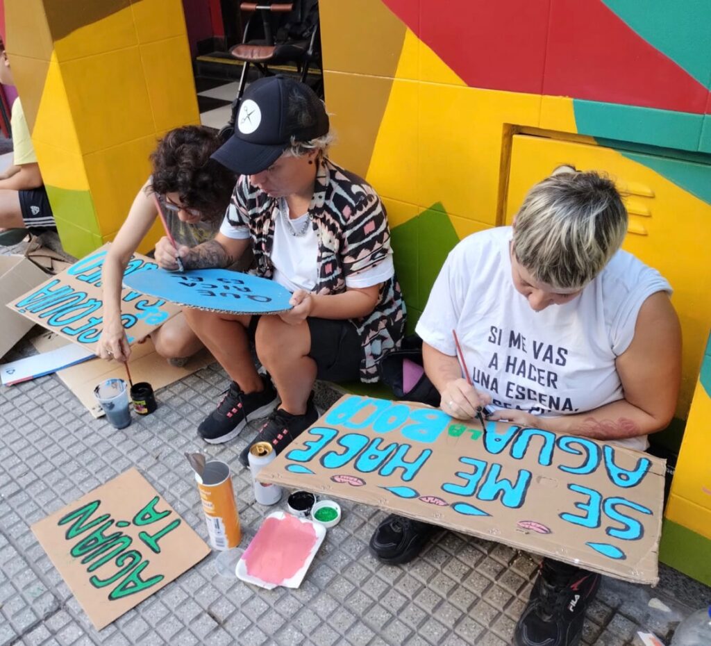 Fotografía donde se ven a tres personas sentadas en la calle pintando carteles en defensa del agua. En los carteles se puede leer: "Aguan-tá", "Se me hace agua la boca", "Sequía - saqueo".