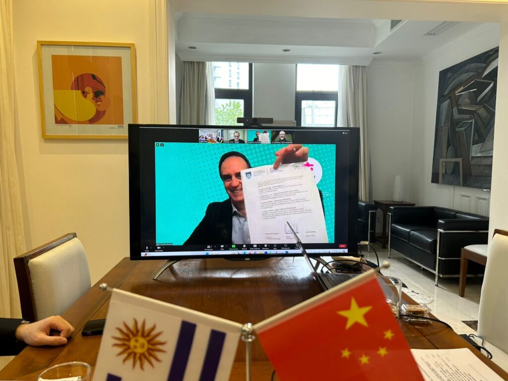 Fotografía donde se ve una mesa con las banderas de Uruguay y China y en el extremo un monitor con la conexión de zoom donde se ve al decano Miranda con el convenio en mano.