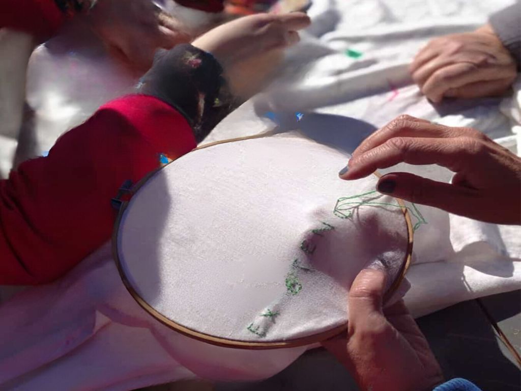 Fotografía que muestra en primer plano un bastidor con una tela blanca colocada y manos bordando letras en color verde.