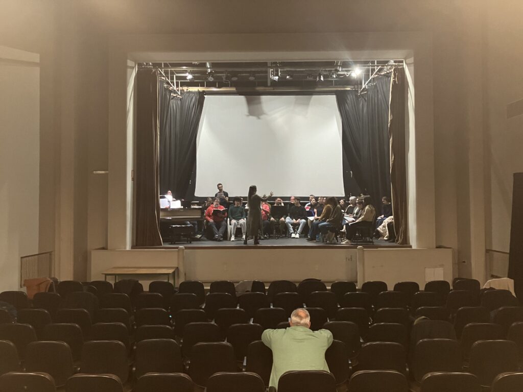 Fotografía del auditorio de Facultad de Artes mirando hacia el escenario. Arriba se ven los integrantes del coro, debajo en las butacas se ve una persona que los observa.