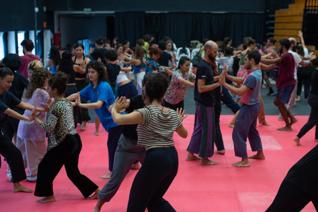 En la imagen se ven muchas personas en parejas, en un salón amplio con piso de tatami, uniéndose palma con palma y en movimiento. 