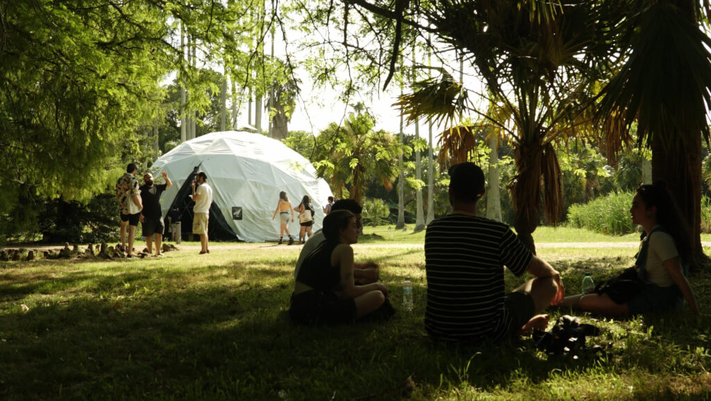 Fotografía donde se ve un espacio de área verde en el jardín botánico, personas sentadas en primer plano y al fondo una especie de domo, carpa blanca